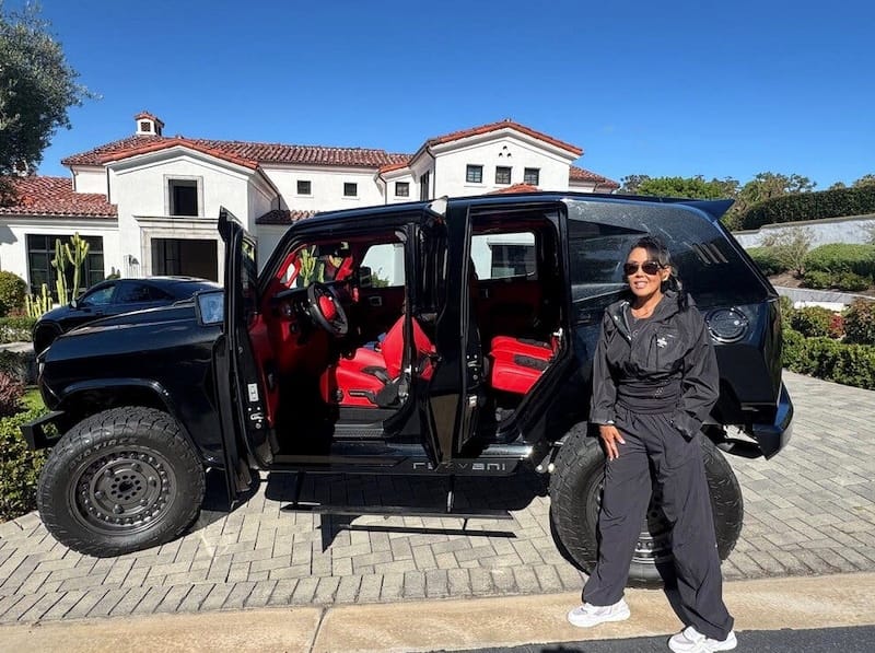 Photo of a woman posed in front of a black, tank-like vehicle. She's wearing sunglasses and a black tracksuit.