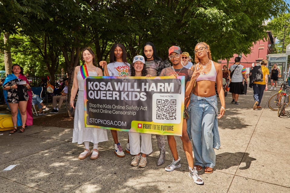 Color photograph of six people standing behind a sign that reads "KOSA WILL HARM QUEER KIDS"